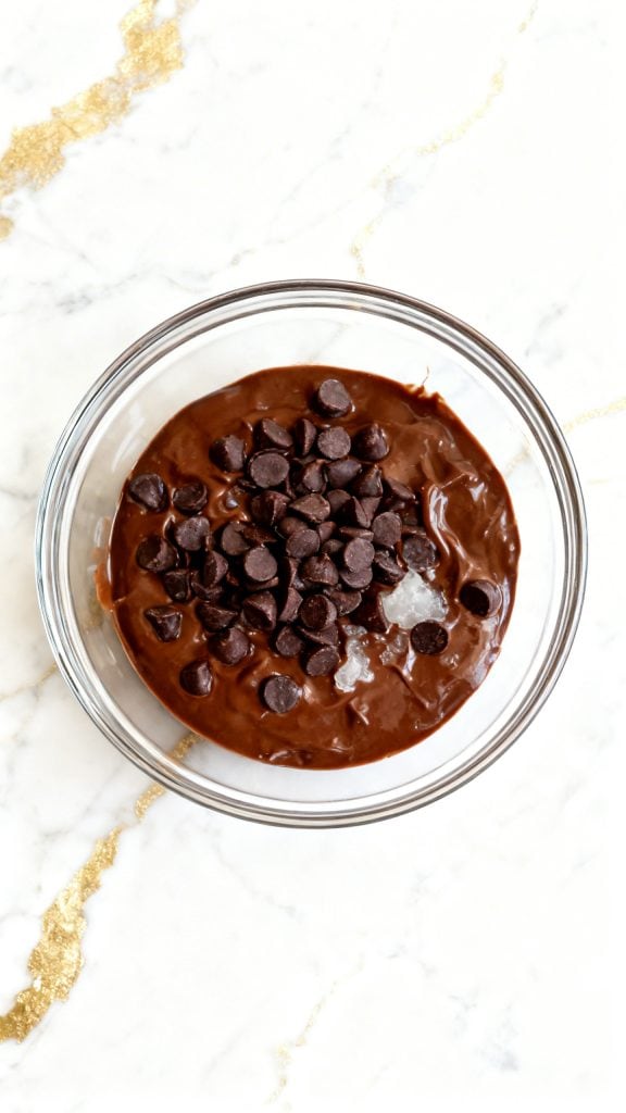 Ultra close-up of fully mixed light brown cocoa batter in a clear glass bowl, thick and smooth consistency with no dry streaks, surface glossy and evenly blended, a straight 90-degree top-down shot with no tilt, captured directly overhead, white marble surface with subtle gold hints
