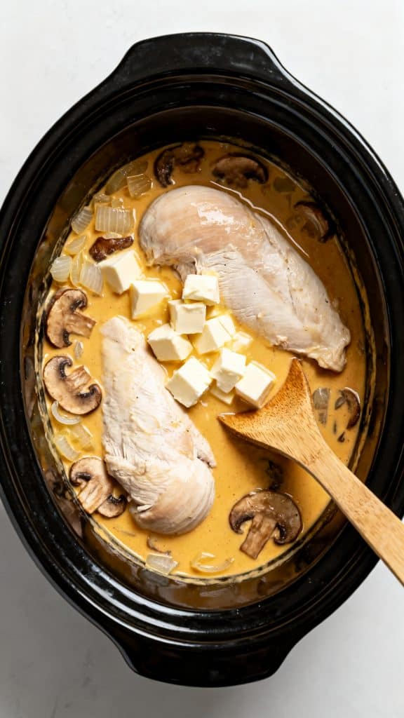 Top-down shot of raw boneless skinless chicken breasts on a large white round plate, Italian dressing seasoning in a small white bowl, sliced unsalted butter on a small white round plate, cream of chicken soup in a white bowl, golden mushroom soup in a white bowl, softened cubed cream cheese on a small white round plate, chicken broth in a clear glass jar, sliced mushrooms in a white bowl, whole onion placed directly on the surface with no holder, uncooked angel hair pasta neatly arranged on a large white round plate, chopped fresh parsley in a small white bowl, salt in a small white bowl, ground pepper in a small white bowl, every ingredient fully visible and neatly spaced apart, main object fully in frame and not cropped out, a straight 90-degree top-down shot with no tilt, captured directly overhead, white marble with hints of gold
